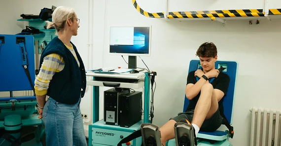 Eine blonde Frau schaut auf einen Monitor. Auf einem Sitz neben ihr sitzt ein junger Mann mit dunklen Haaren, ein Bein auf dem Boden, eins auf einem Pedal, die Arme vor der Brust gekreuzt.