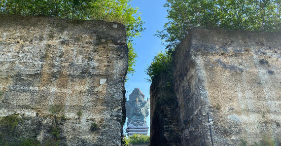 Die Garuda-Wisnu-Kencana-Statue ist in der Ferne im Hintergrund zu erkennen. Sie ist sehr groß und thront auf der gesamten Dachfläche eines großen Gebäudes. Die Statue ist durch den Spalt zwischen einer Felsformation mit sekrechten Wänden zu erkennen.