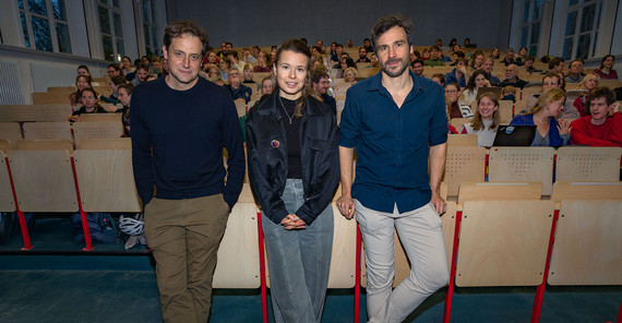 Drei Personen stehen mit dem Rücken zum Auditorium eines gut gefüllten Hörsaal und posieren für das Foto.