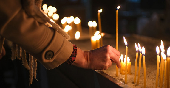 A hand can be seen placing a lit thin candle next to other candles that are already burning. The warm light of the candlelight illuminates a scene that would otherwise be dark.