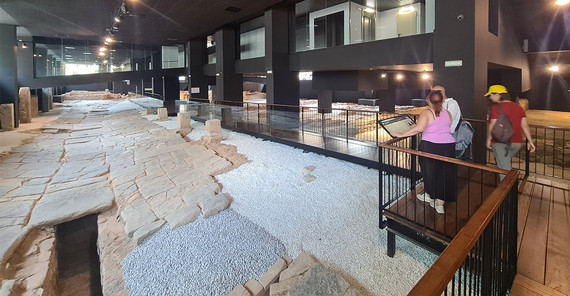 Ein freigelegter römischer Straßenzug unter dem Bodenniveau der heutigen Stadt. Der Straßenzug ist überdacht und von künstlichem Licht beschienen. Menschen können ihn nicht betreten, für sie sind Plattformen mit Absperrungen installiert.