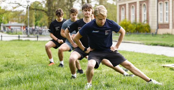 Fünf junge Männer machen auf einer Wiese Dehnungsübungen.
