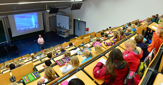 Hörsaal, Blick von oben in Richtung Professor. Kinder haben ihre Hefte und Federmappen vor sich auf den Tischen und hören aufmerksam zu.