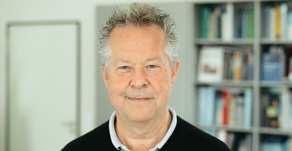 Ein Mann mit grauen Haaren in einem schwarzen Pullover. Im Hintergrund sieht man unscharf ein Bücherregal.