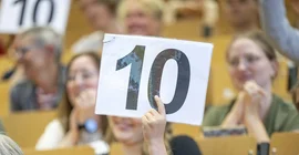 Hand, die ein Schild mit einer 10 darauf hochhält, im Hintergrund Menschen in einem Hörsaal.