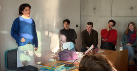 Person hält einen Vortrag. Teil des Publikums mit freundlichen Gesichtern erkennbar.