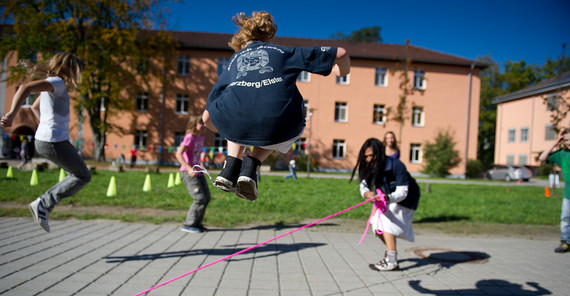 Kinder springen über ein langes Seil, gedreht von einem Mädchen in der Mitte. Im Hintegrund Uni-Gebäude.