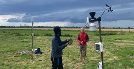 Zwei Personen stehen auf einem Feld an einem wissenschaftlichen Messgerät, im Hintergrund dunkle Gewitterwolken