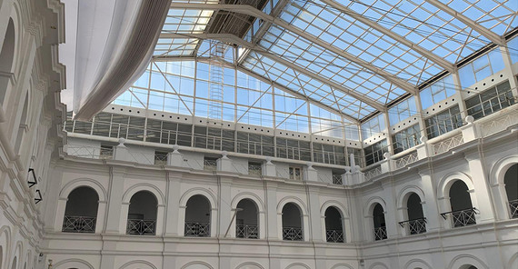 Ein Blick nach oben in das gläserne Dach des Archäologischen Museums durch welches blauer Himmel zu erkennen ist. Die Metall-Glaskonstruktion wird getragen von weißen Wänden, die rhythmisch auf mehreren Etagen durch Torbögen unterbrochen sind.