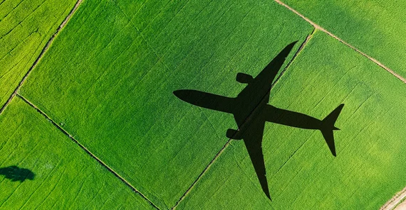 Vogelperspektive auf grüne Felder. Auf ihnen bildet sich der Schatten eines darüber fliegenden Flugzeuges ab.